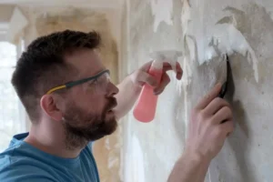 Man scraping textured paint off a wall during a how to remove texture from walls home remodeling project.