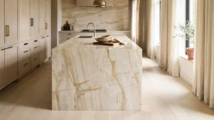 Modern kitchen with a waterfall edge quartzite island in warm cream and gold veining, matching stone backsplash, and light wood cabinetry
