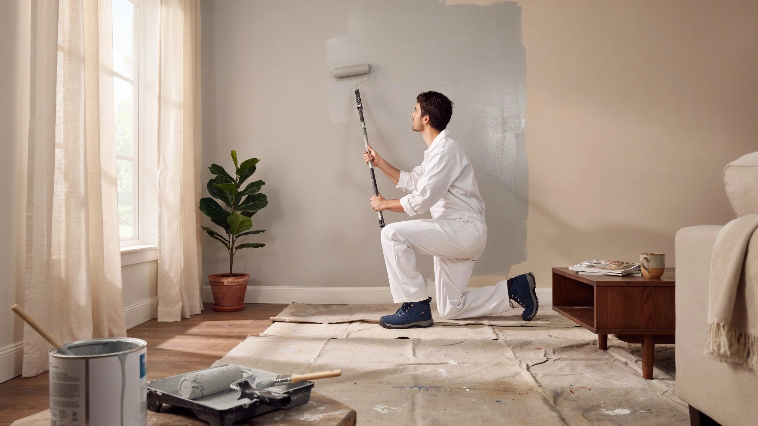 Professional painter in white coveralls rolling gray paint onto a living room wall during a 1000 sq ft interior painting project
